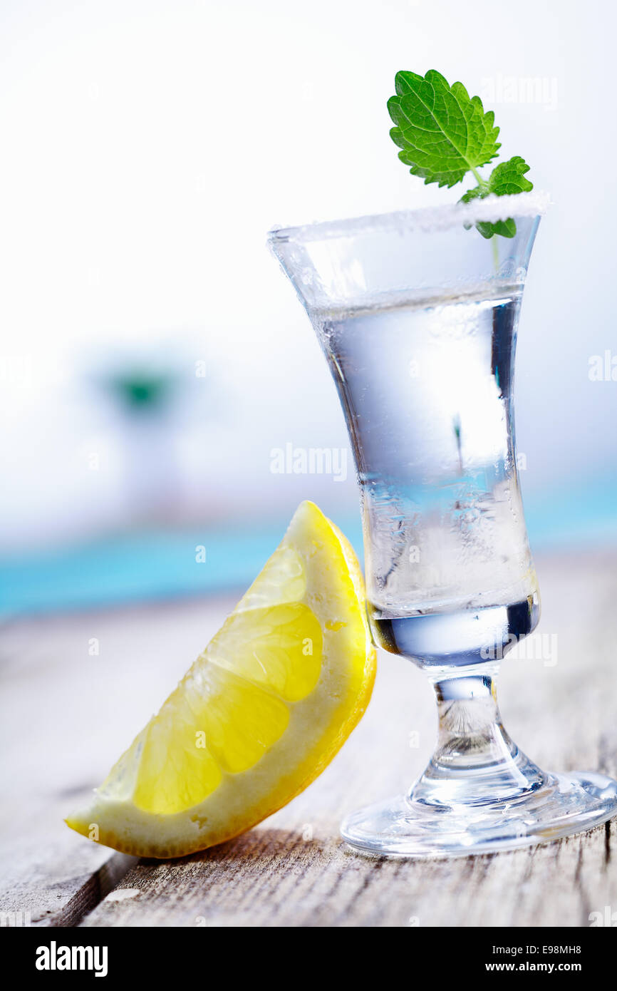 Cold vodka in a frosted shot glass served with lemon chaser alongside a blue pool in summer Stock Photo