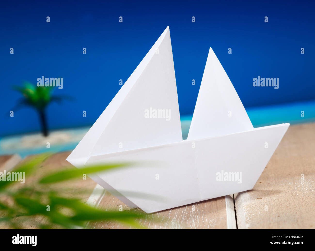a paper boat on a wooden table in front of a beach with a palm on an idyllic place. Maybe for holiday vacation or wellness concepts Stock Photo