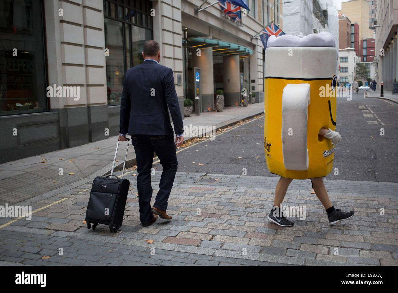Dressed up as a glass of beer in the City of London, UK. Weird street scene as a man in a suit passes this large scale glass. Stock Photo