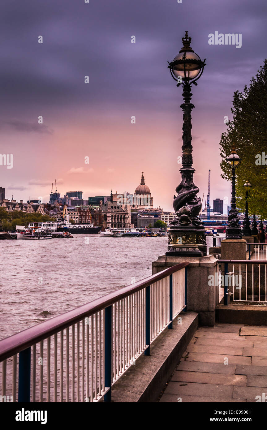 A railing at the waters edge of the Thames River leads the the icon street lamps which are glowing in the evening light. Stock Photo