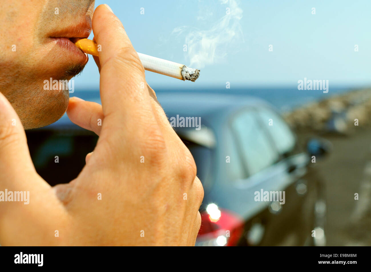closeup of a man with a burning cigarette in his mouth while is waiting besides a car parked next to a no traffic road Stock Photo