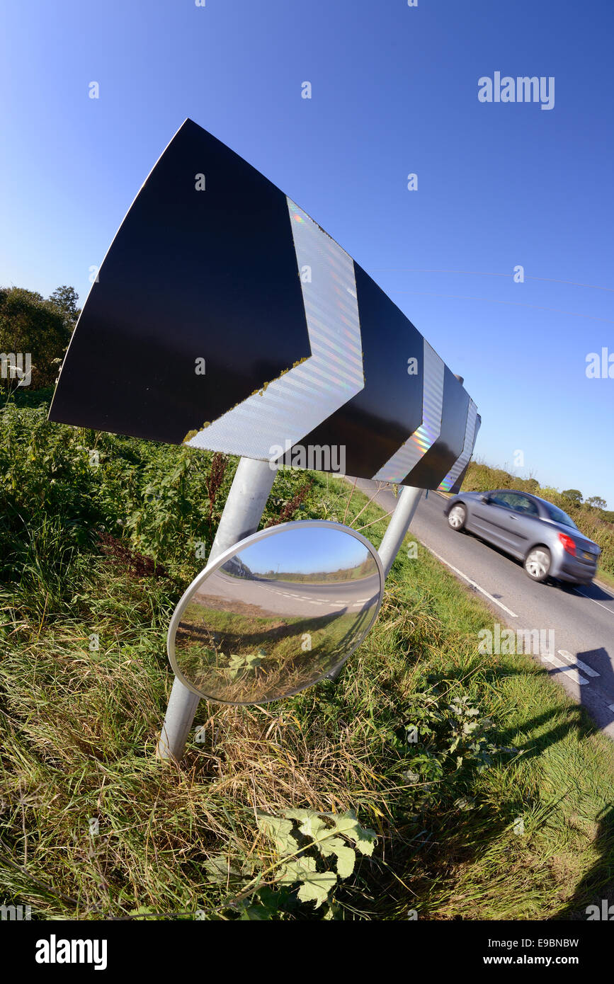 car passing warning sign of sharp bend and circular safety mirror to give drivers emerging from junction view of road uk Stock Photo