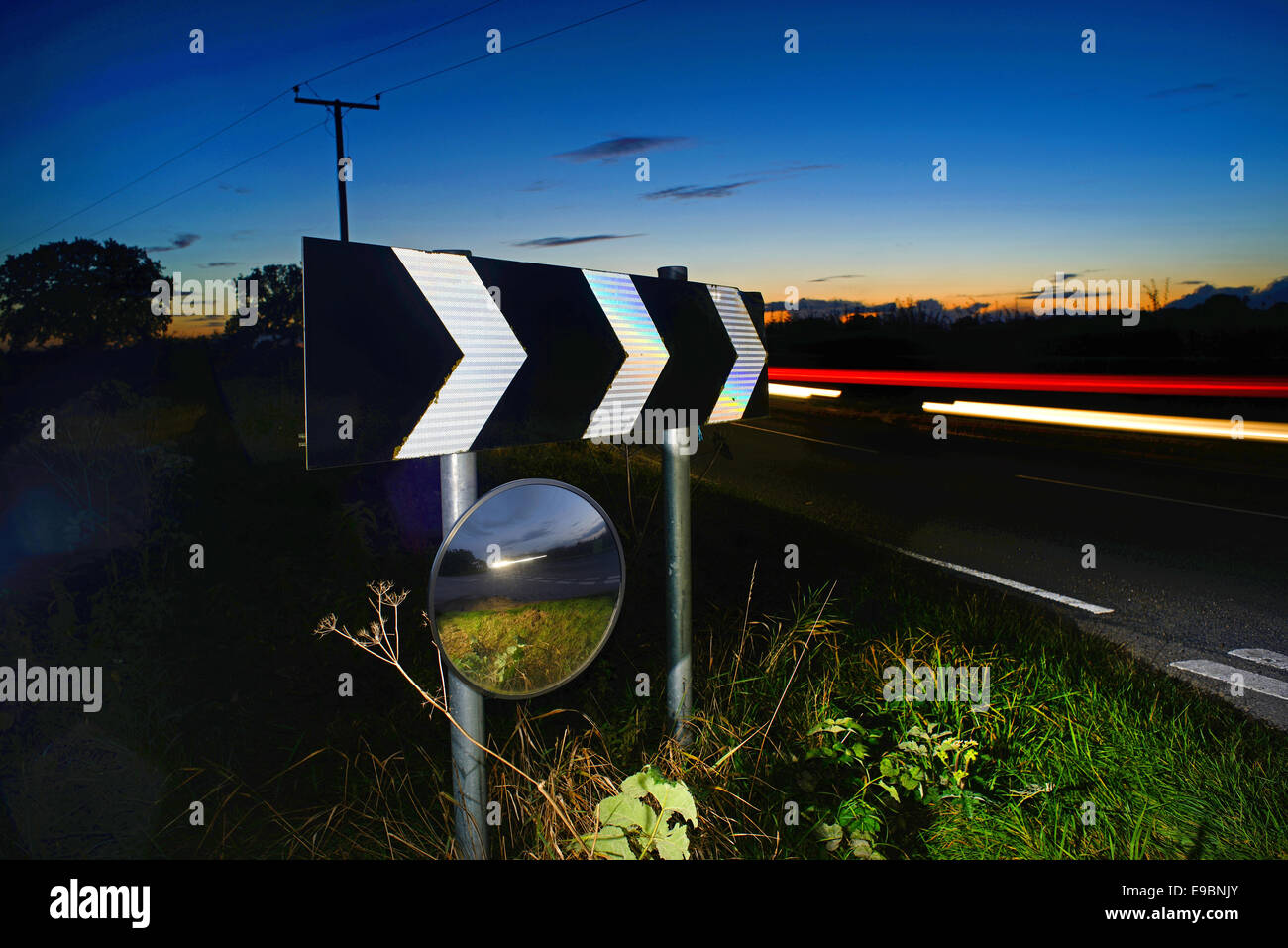 traffic at twilight passing warning sign of sharp bend and circular safety mirror to give drivers  view of road uk Stock Photo