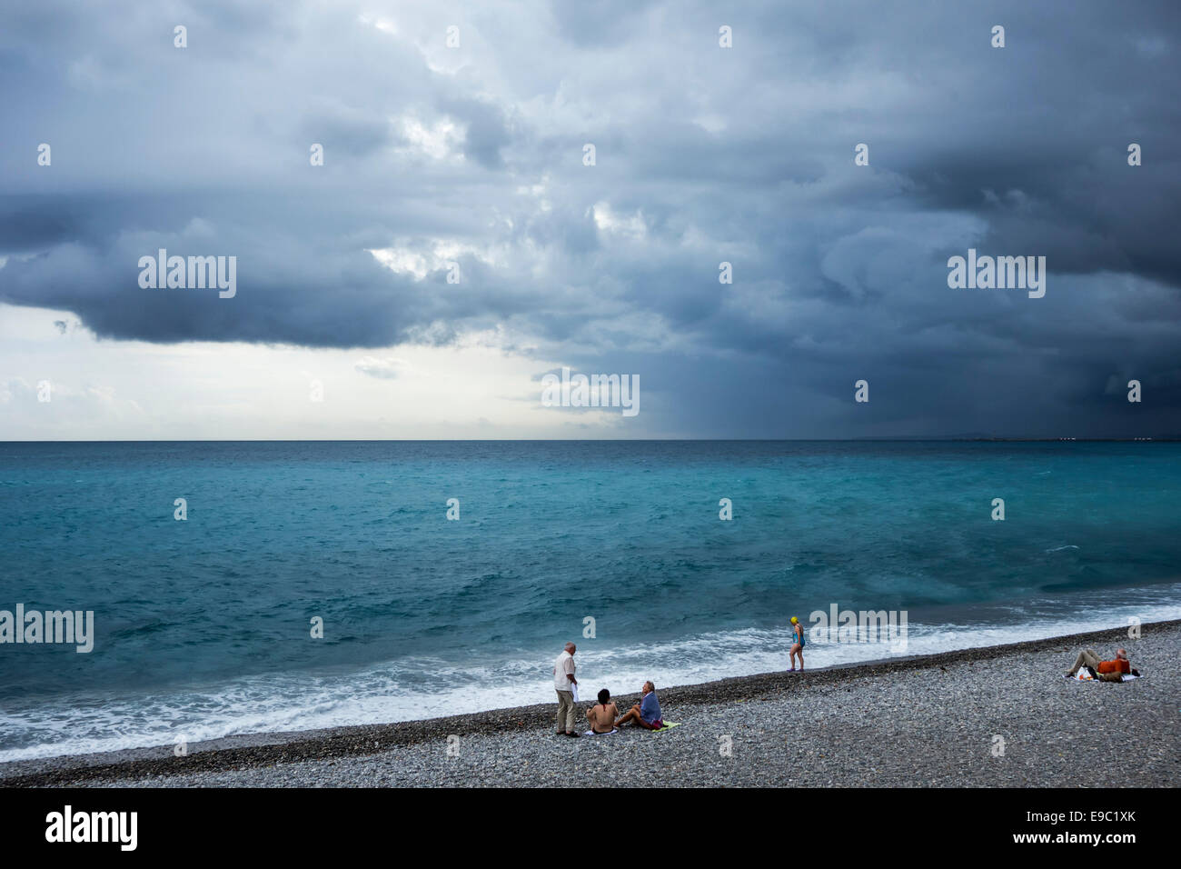 Female swimmers and sunbathers sitting on shingle beach during bad weather with rainstorm approaching over the sea Stock Photo