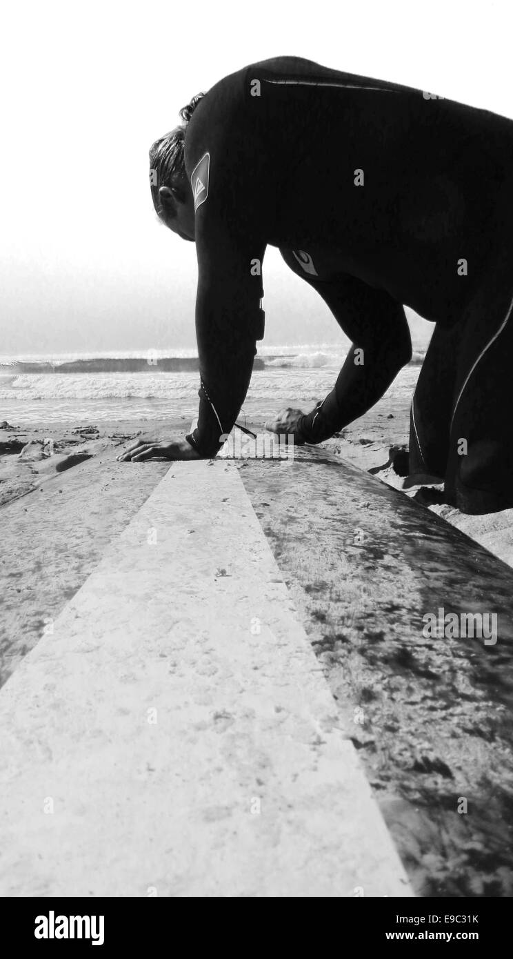 Surfer guy waxing up his long board with ocean in the background in Black and white Stock Photo