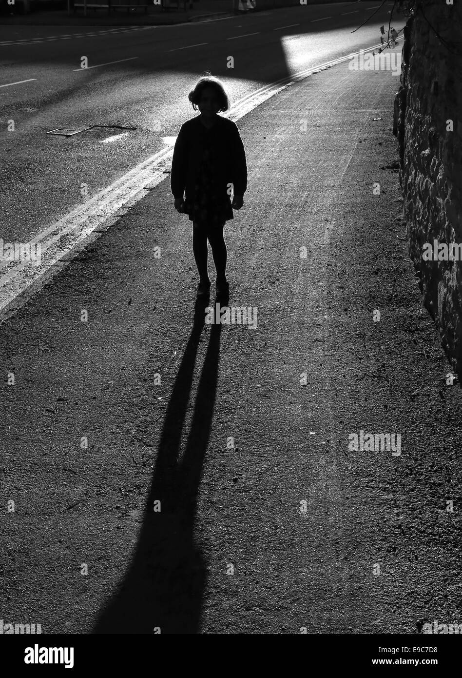 Young girl standing alone silhouetted by striking bright low evening sun on empty  pavement, with a long shadow and backlit hair Stock Photo