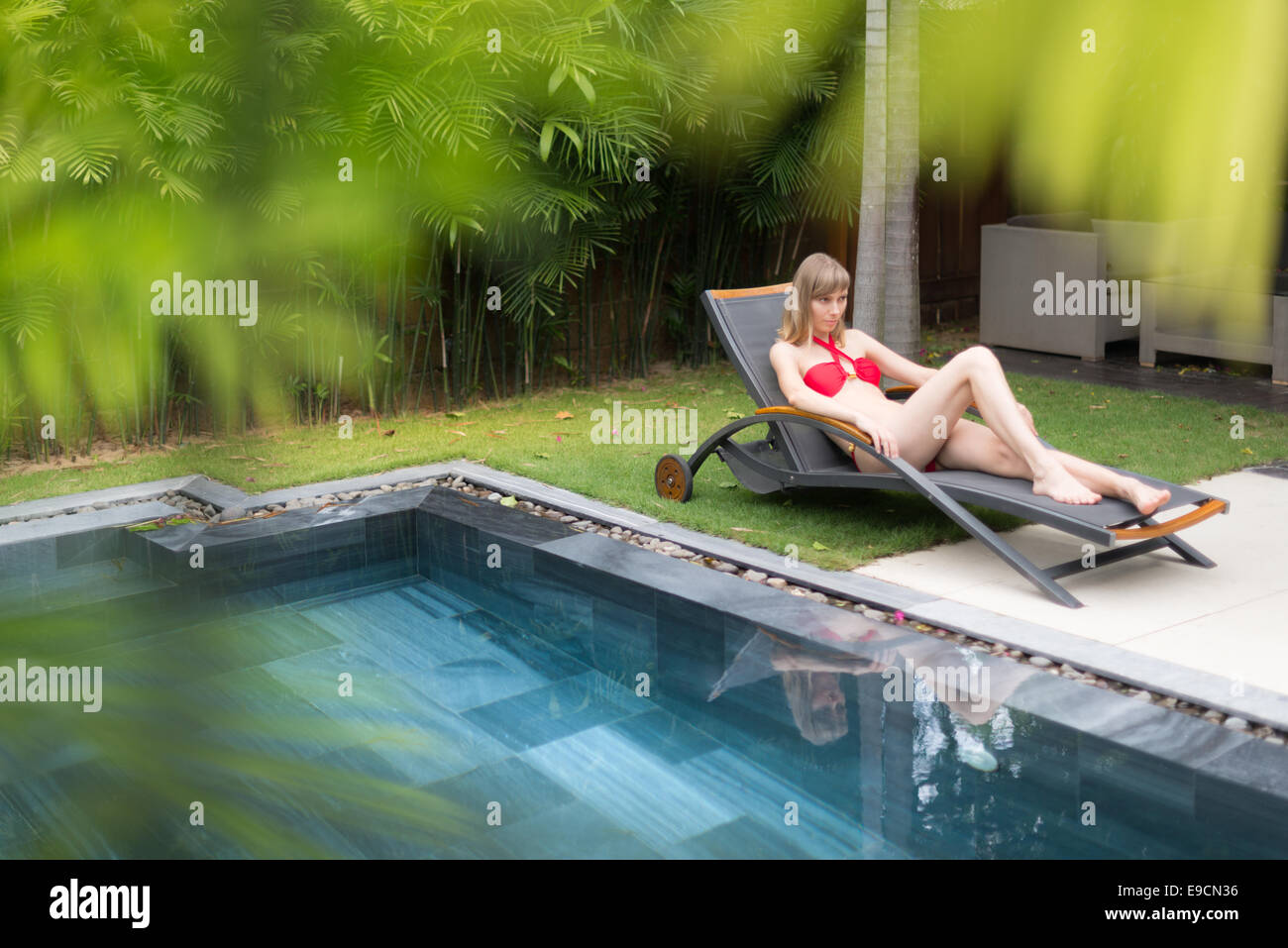 Young woman in red swimsuit relaxing on chaise longue near pool. Blurred palm tree leaves in foreground. Stock Photo