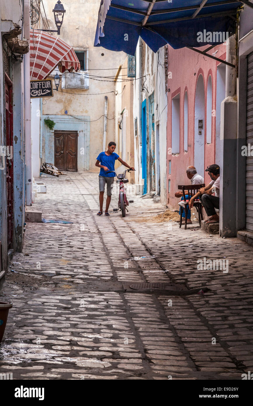 Young man walks his moped down a narrow cobbled sidestreet in the Medina in Sousse,Tunisia while two others chat on a doorstep. Stock Photo