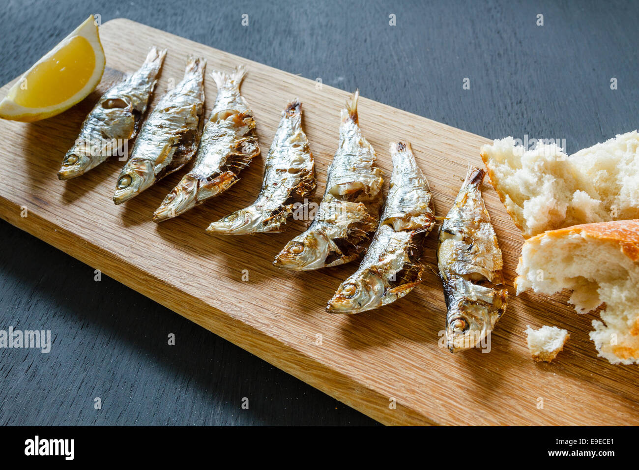 Fresh grilled sprats on a wooden chopping board with a lemon wedge and chunks of bread Stock Photo