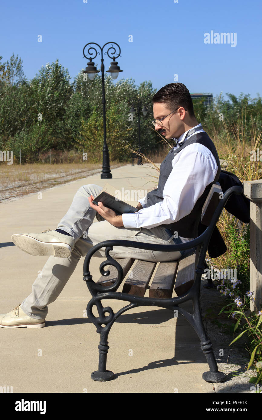 Pensive man with mustache sitting on bench reading book wearing glasses relaxed on sunny day Stock Photo
