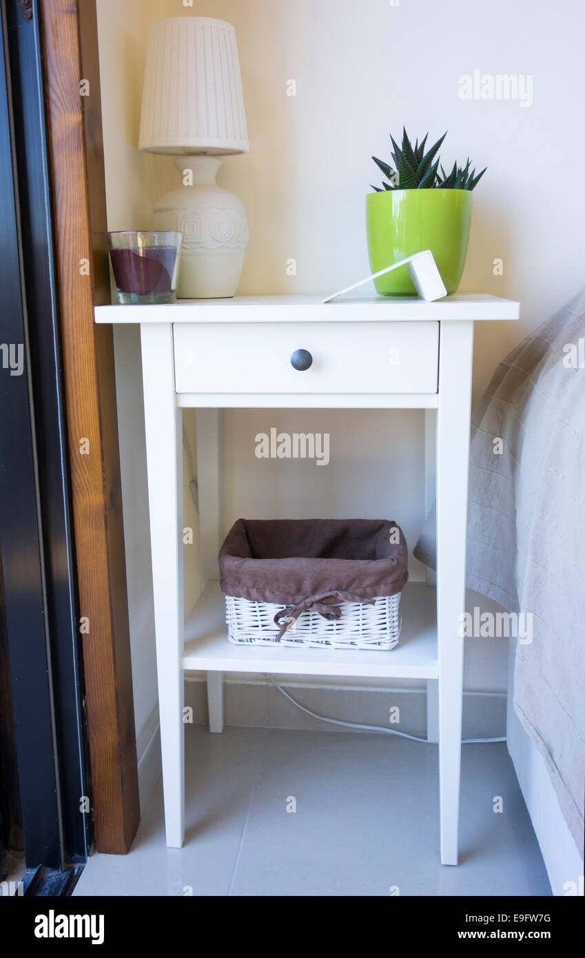 White bedside table with table lamp, candle, flower pot, basket and