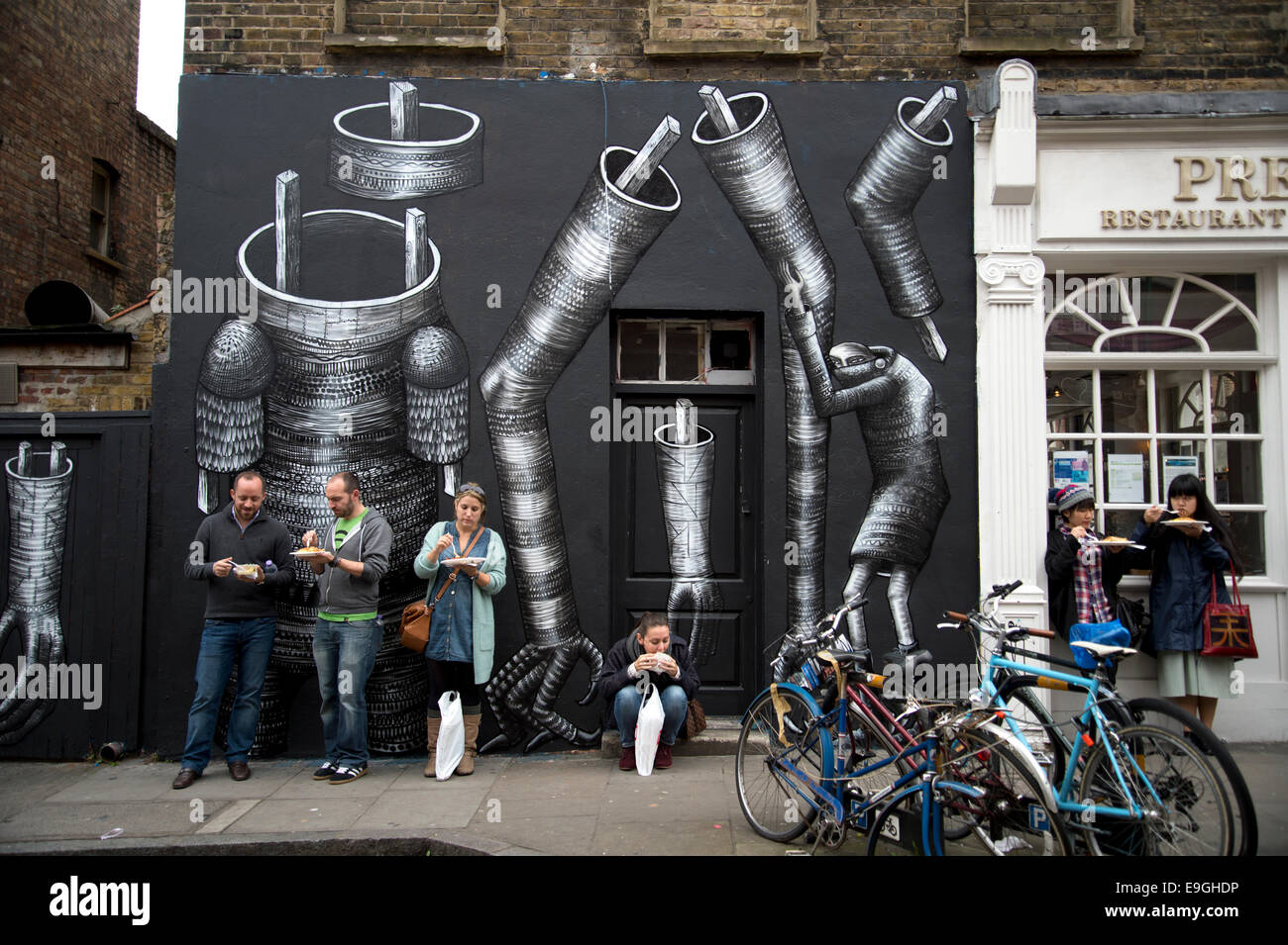 Brick Lane. Sunday lunchtime. People eat on the street next to a piece of street art. Stock Photo