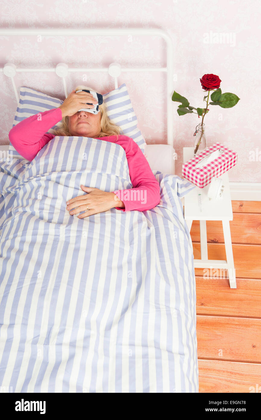 Sick woman in pink pajama with migraine, tissues and a rose lying in bed with washcloth Stock