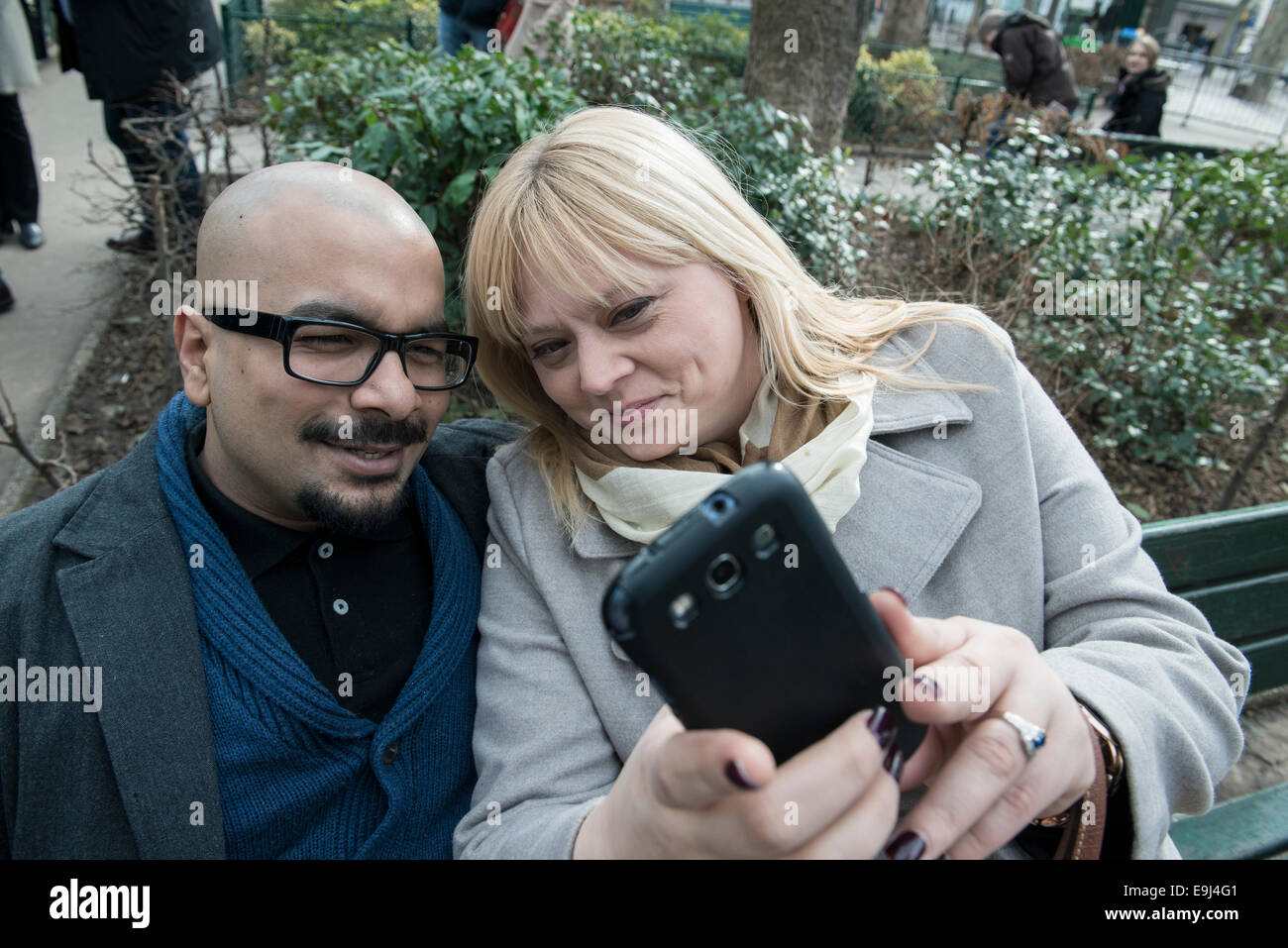a real authentic couple sit on a bench in paris and take a selfie on a smart phone while looking happy Stock Photo