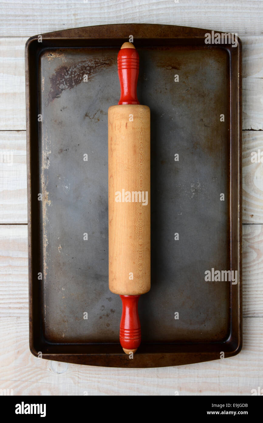 High angle closeup of an old wooden rolling pin with red handles on a well used cookie sheet. The pan is on a rustic table Stock Photo