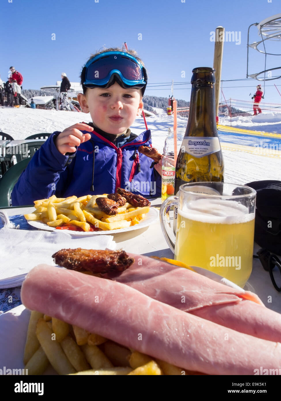 small boy in ski clothes eating at an outdoor table next to a ski piste with beer and food in foreground Stock Photo