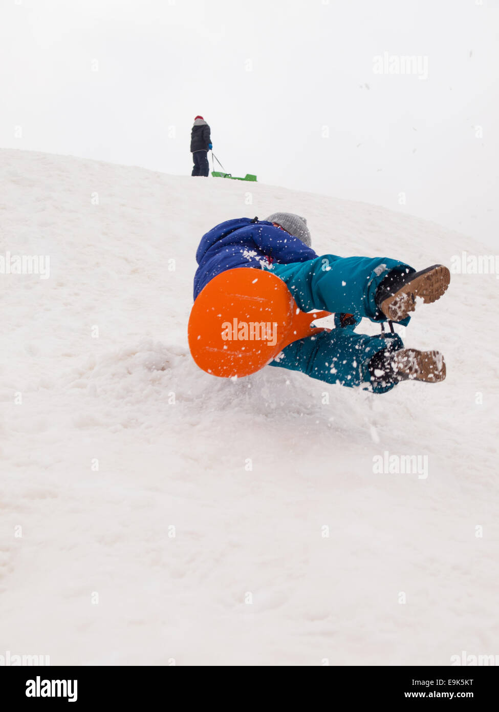 small boy sledging fast down a steep hill with child standing at top of ...