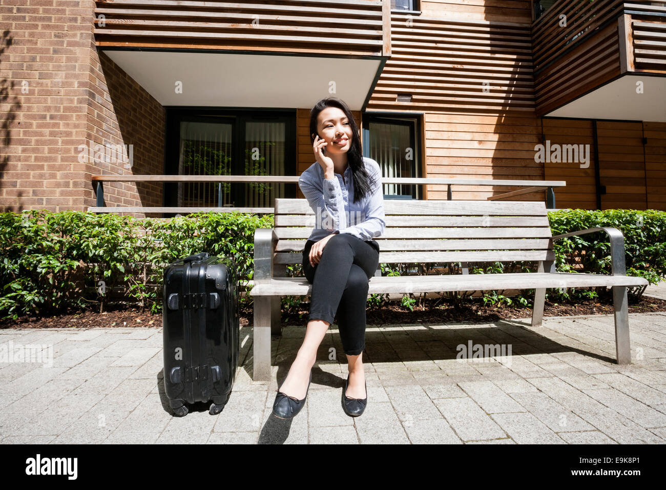 Full length of businesswoman answering cell phone while sitting by luggage on bench against building Stock Photo