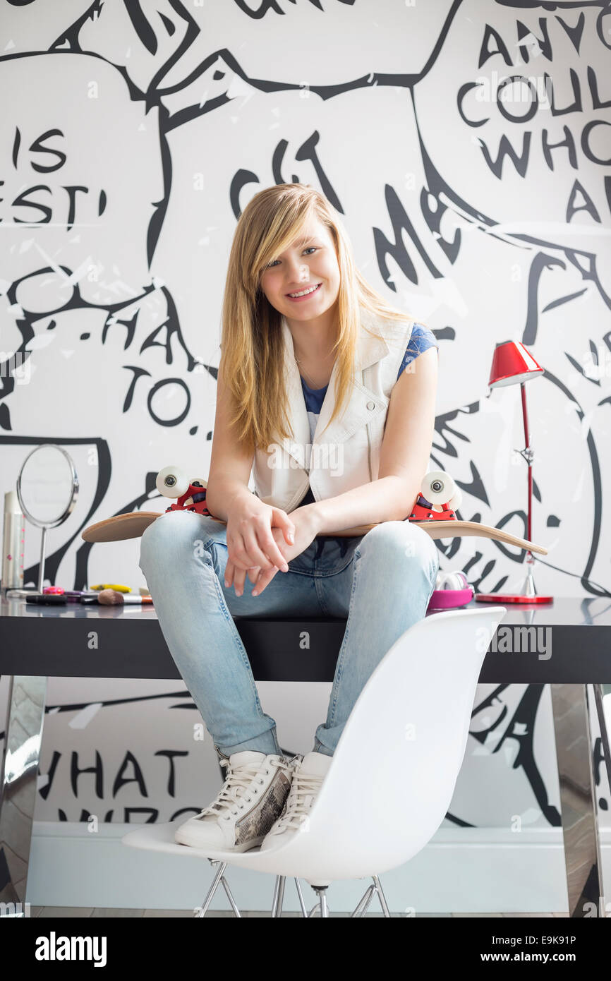 Full-length portrait of happy teenage girl with skateboard sitting on study table at home Stock Photo