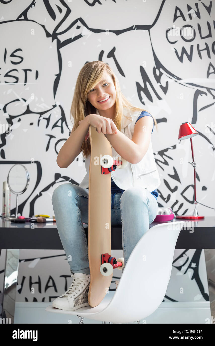 Full-length portrait of happy teenage girl with skateboard sitting on study table at home Stock Photo