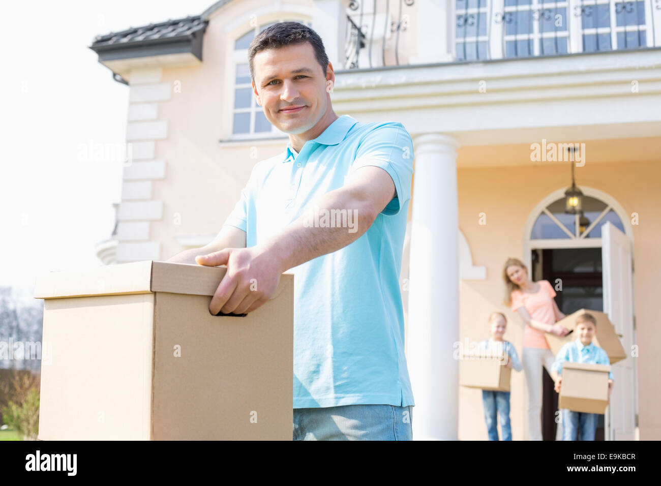 Portrait of confident man carrying cardboard box while moving house with family in background Stock Photo