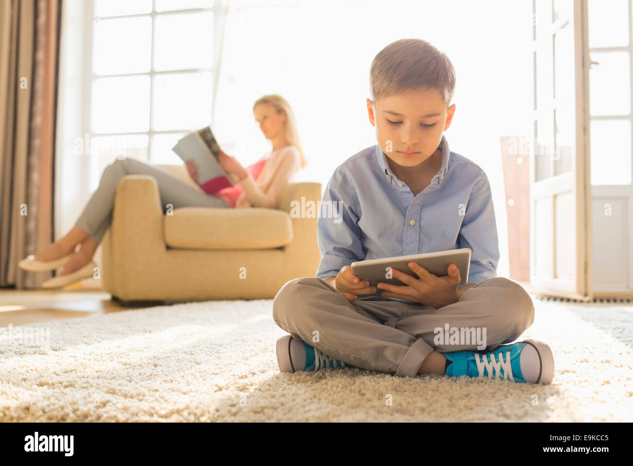 Boy using digital tablet on floor with mother reading magazine in background Stock Photo