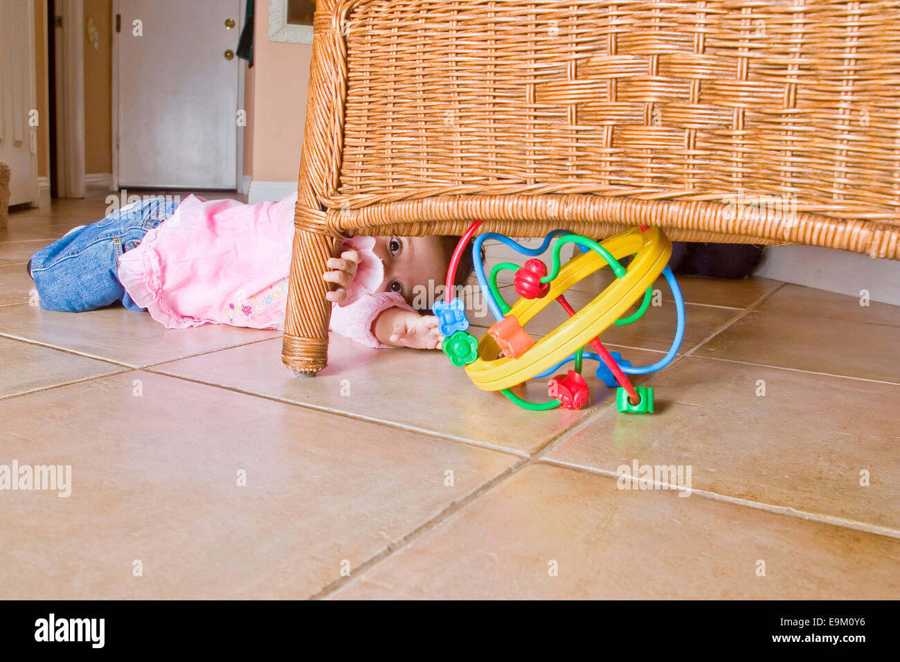 9-month-old Colombian baby girl crawls toward brightly colored toy hidden under rattan chair at home. MR  © Myrleen Pearson Stock Photo