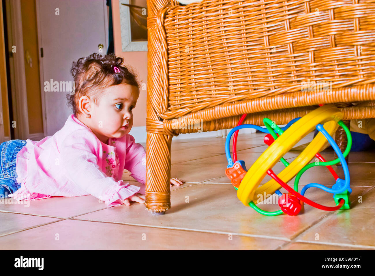 9-month-old Colombian baby girl crawls toward bright colored toy hidden under rattan chair at home. MR  © Myrleen Pearson Stock Photo