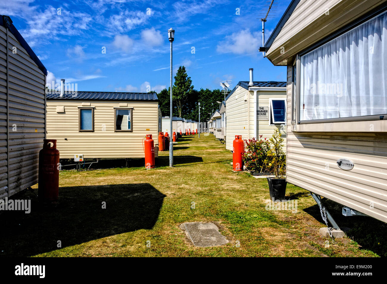 Red gas cylinders stand outside a row of neatly parked cream holiday caravans on either side of a wide path of brown mown grass Stock Photo