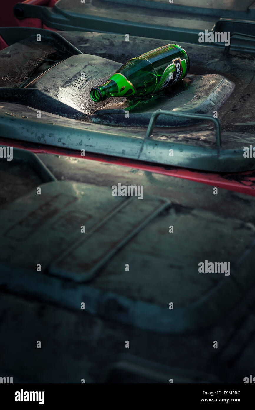 Green glass cider bottle resting on the top of plastic wheelie rubbish bins. Stock Photo