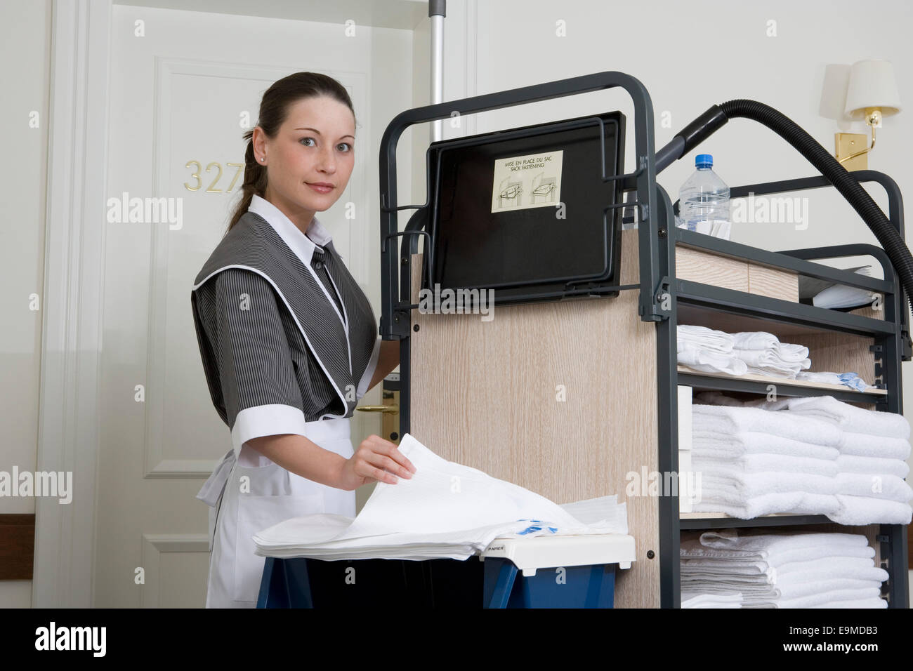 Uniformed maid standing next to push cart in hotel corridor Stock Photo