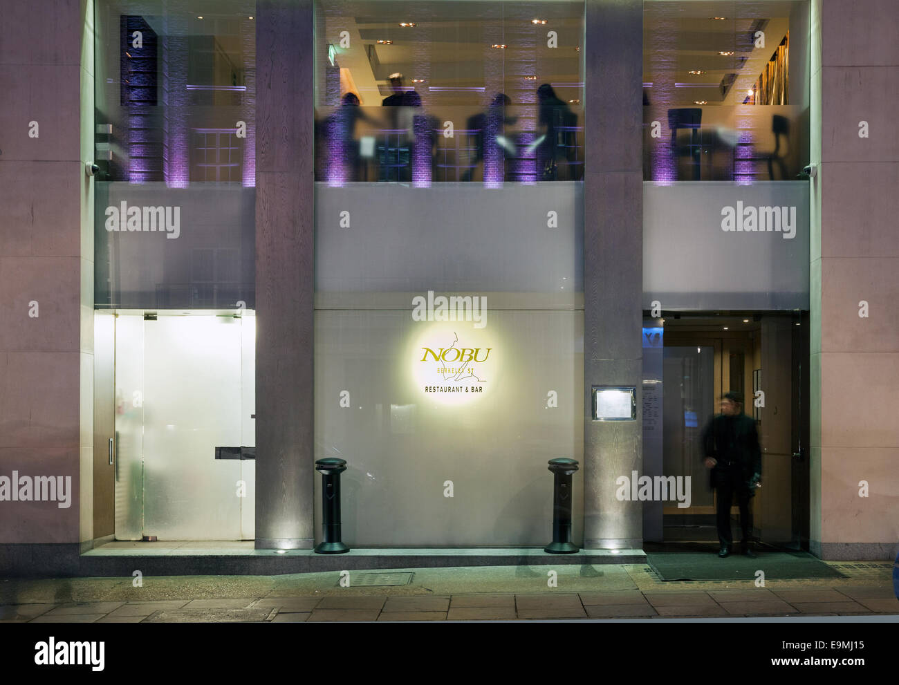 Nobu restaurant   frontage illuminated at night, with diners eating at  the windows and doorman at the entrance Stock Photo