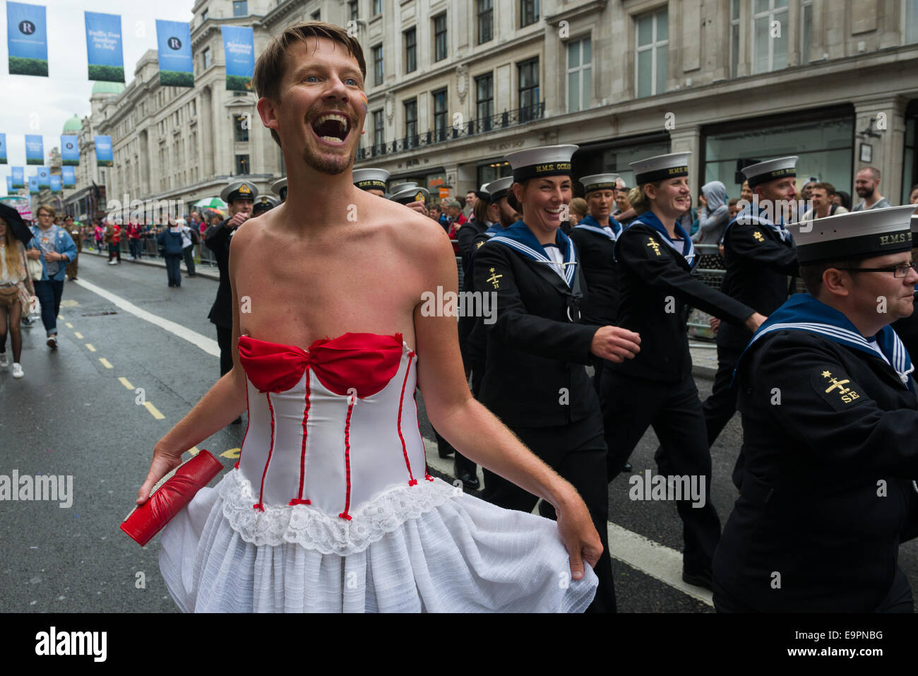 Man wearing a white dress posing in front of members of the Royal Navy marching in the  Pride in London parade 2014, London, England Stock Photo