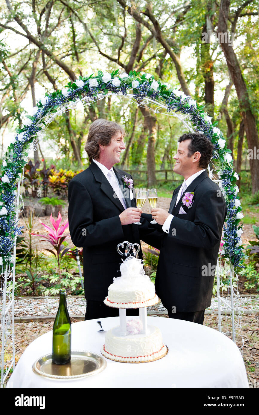 Happy handsome gay couple toasting with champagne under floral wedding arch. Wedding cake with two grooms in foreground. Stock Photo