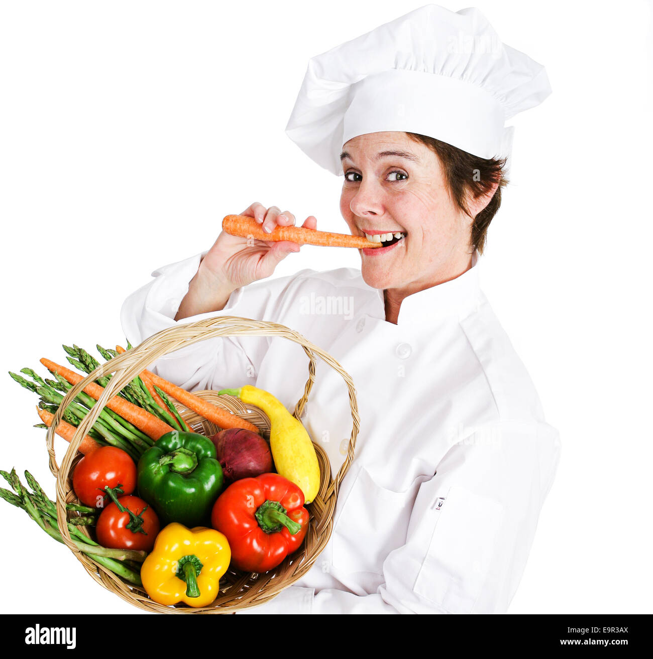 Happy female chef holding a basket of fresh, organic, locally grown produce and eating a raw carrot.  Isolated on white backgrou Stock Photo