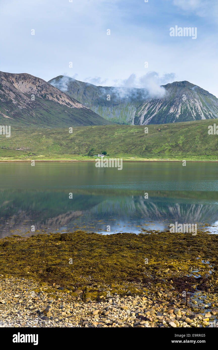 Small white solitary crofters cottage nestling below mountain range reflected in waters of the loch on Isle of Skye in the Highl Stock Photo