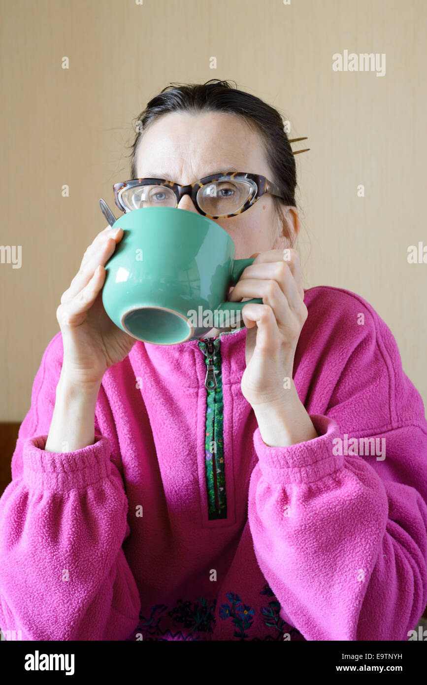 A mature woman with glasses wearing a pink pull-over and drinking coffee in a green cup at morning for breakfast Stock Photo