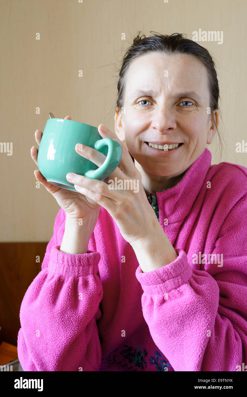 A smiling mature woman wearing a pink pull-over and drinking coffee in a green cup at morning for breakfast Stock Photo