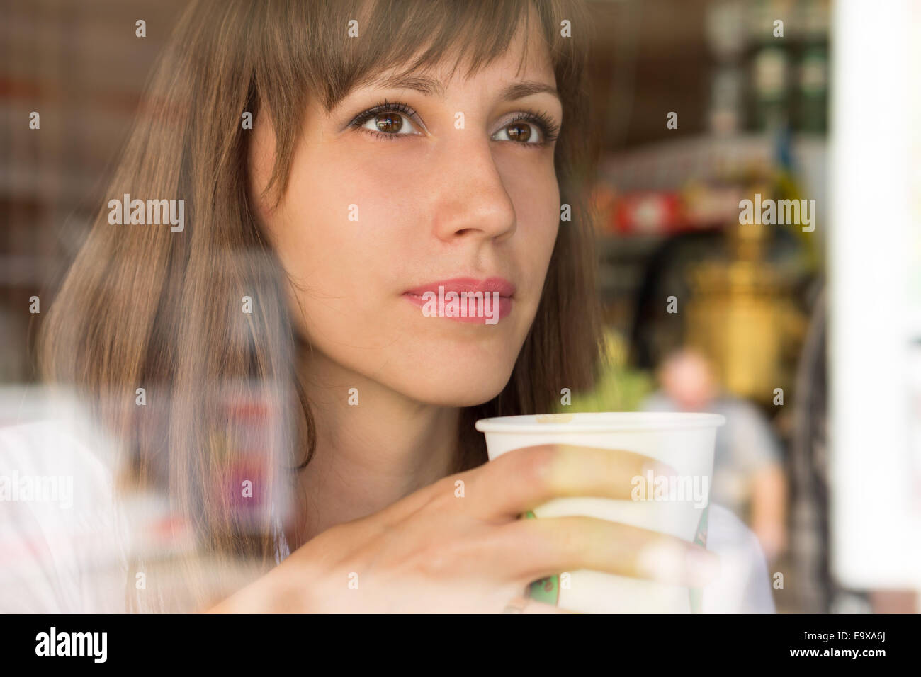 Young thinking woman with paper cup of coffee or tea looking through ...