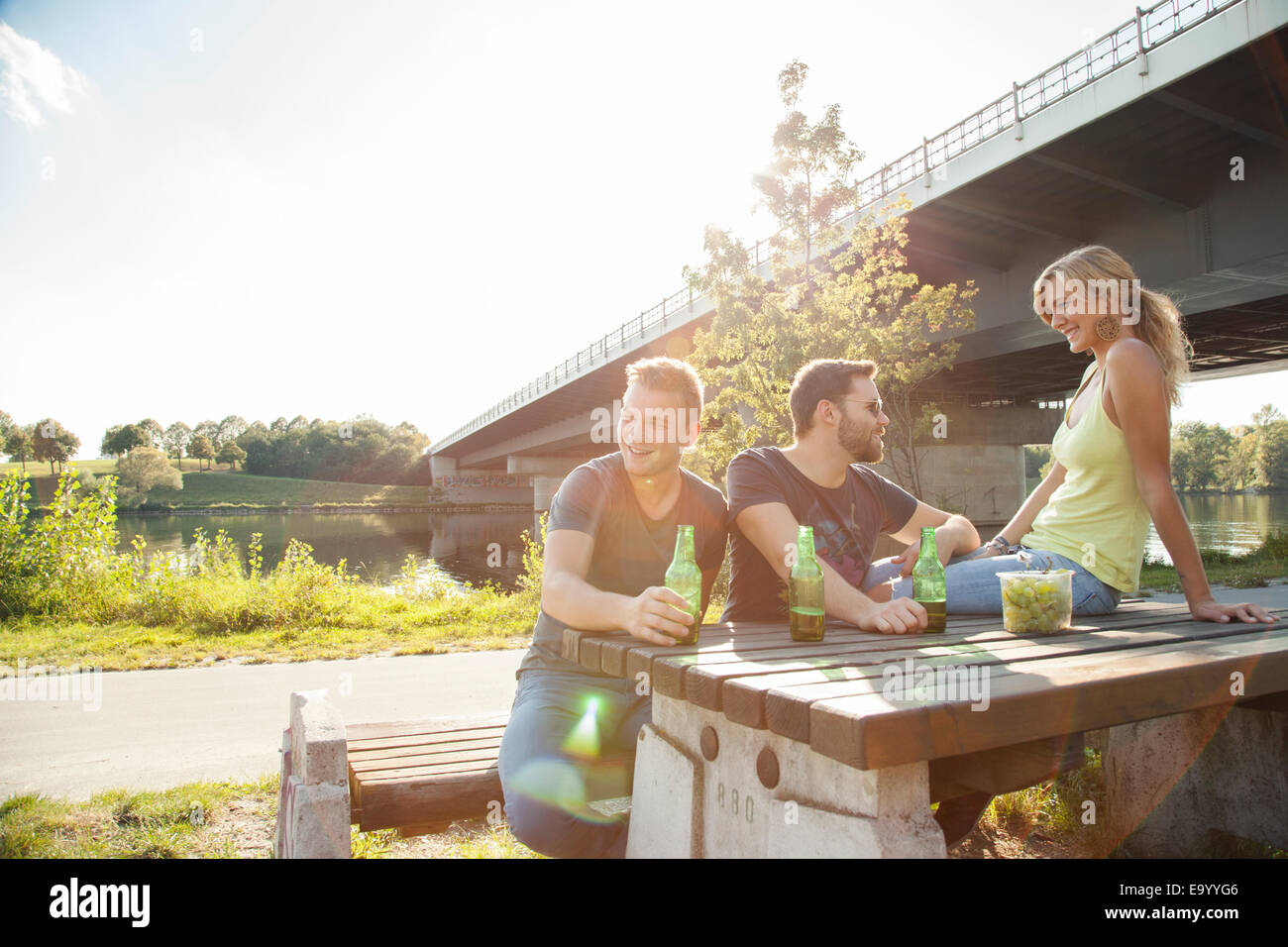 Three young friends drinking beer on riverside picnic bench Stock Photo