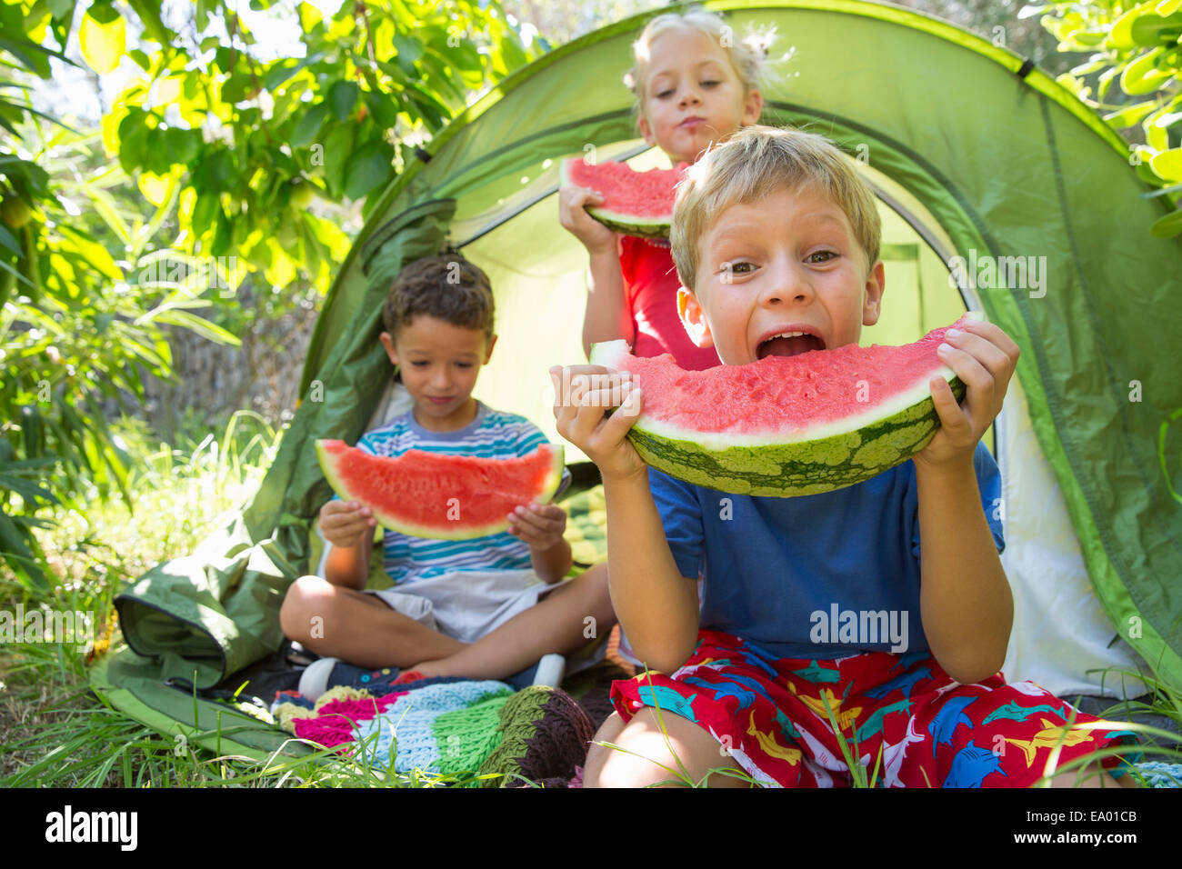 Three children eating large watermelon slices in garden tent Stock Photo