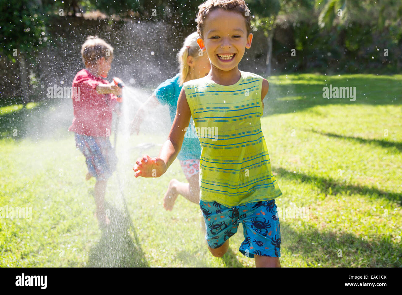 Three children chasing each other in garden with water sprinkler Stock Photo