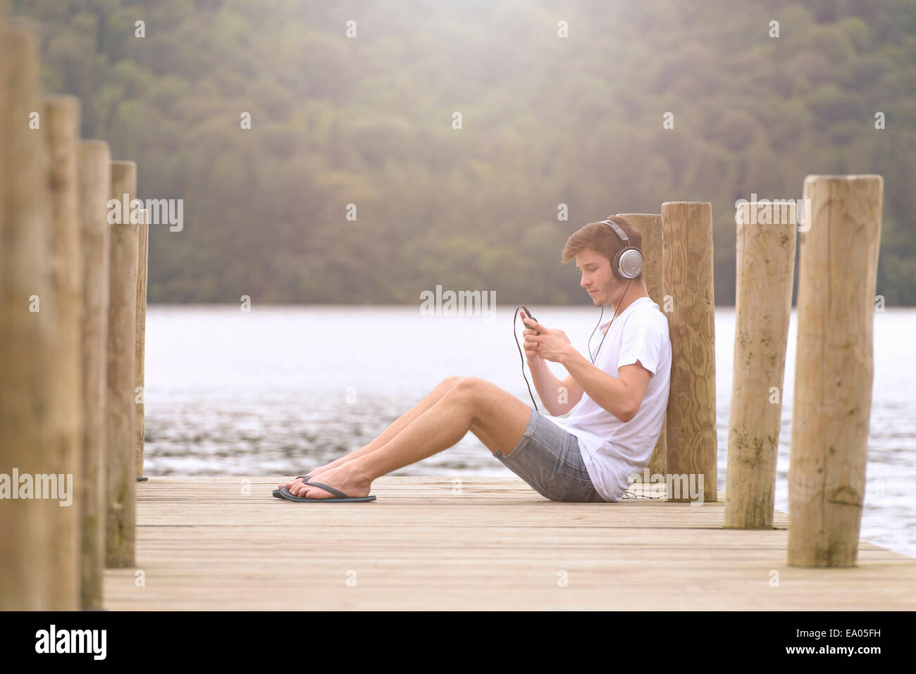 Young man wearing headphones and using mp3 player on jetty over lake Stock Photo