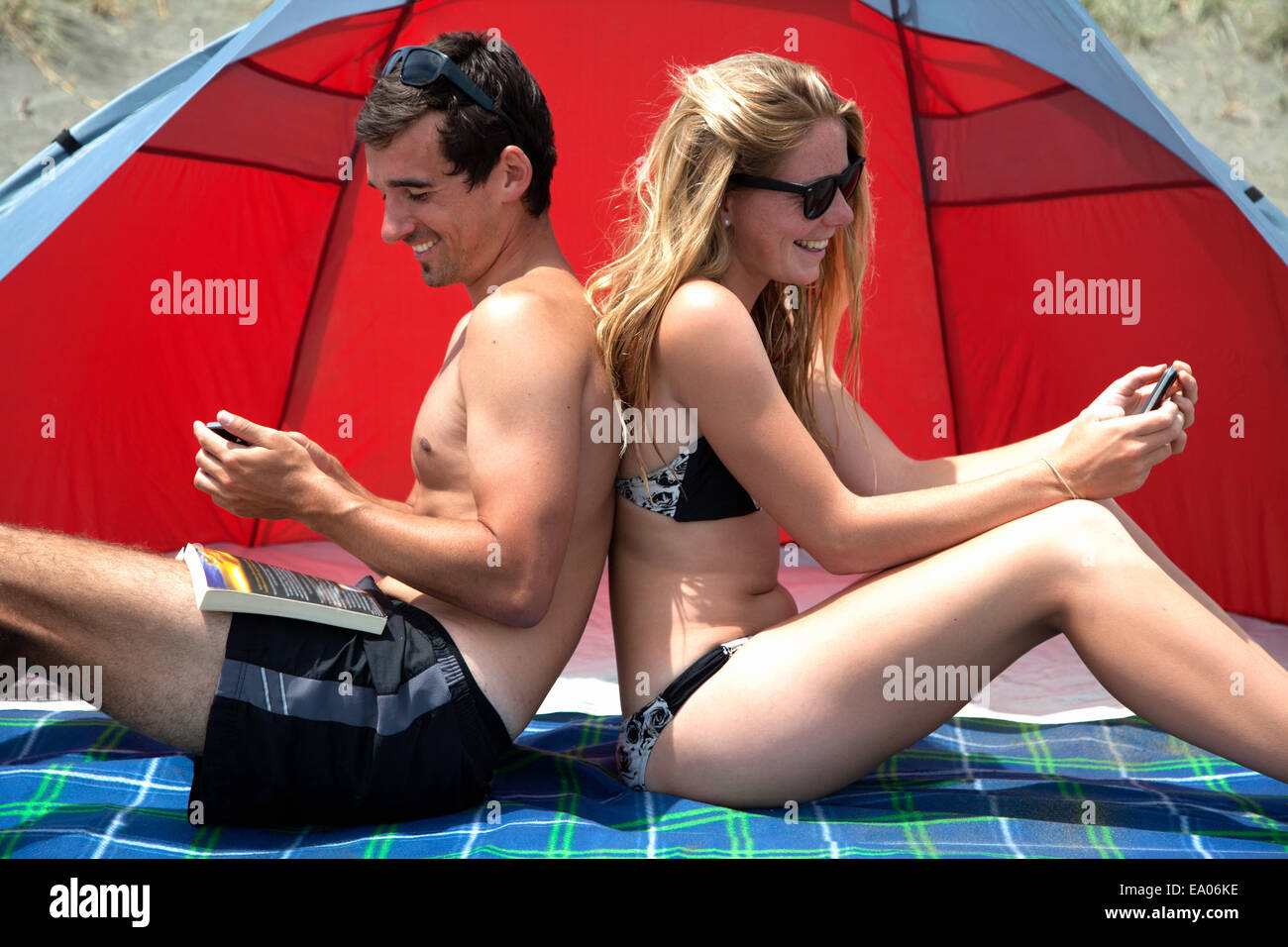 Young couple sitting back to back on beach towel, using smartphones Stock Photo