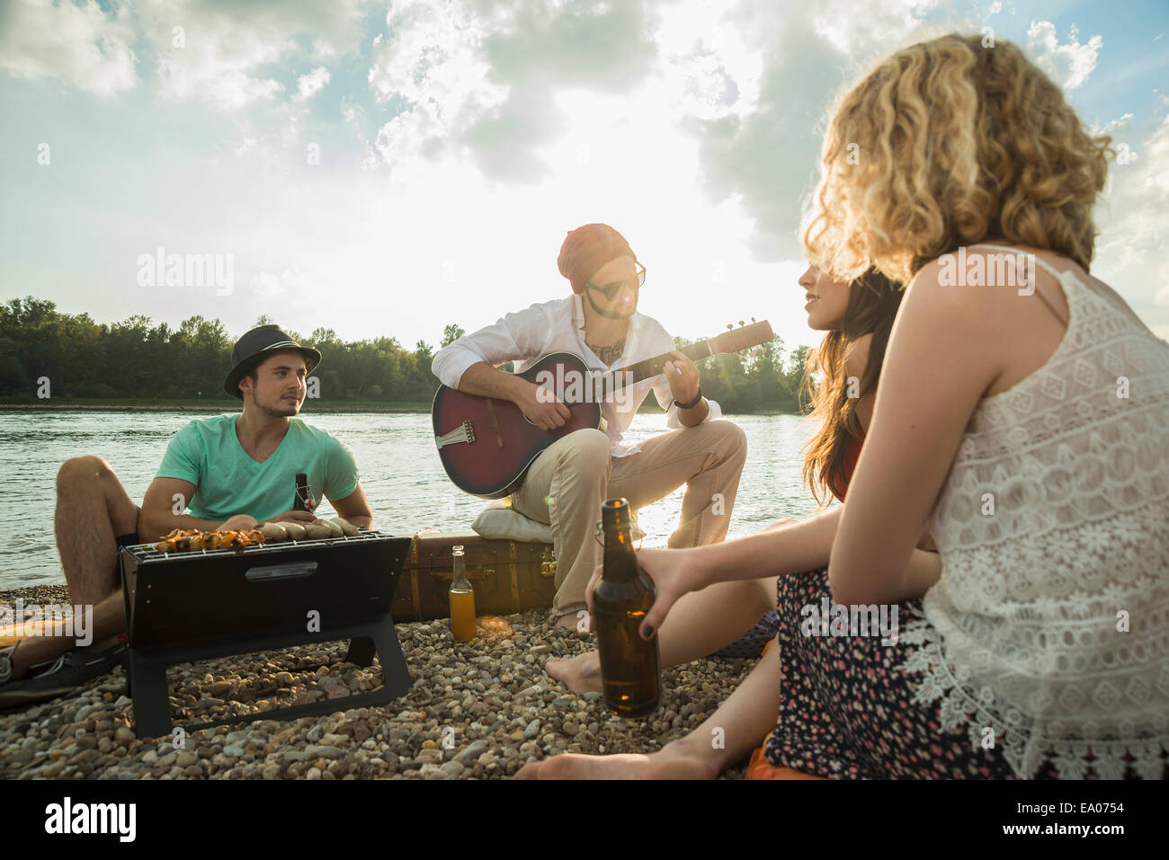 Young man sitting by lake with friends playing guitar Stock Photo