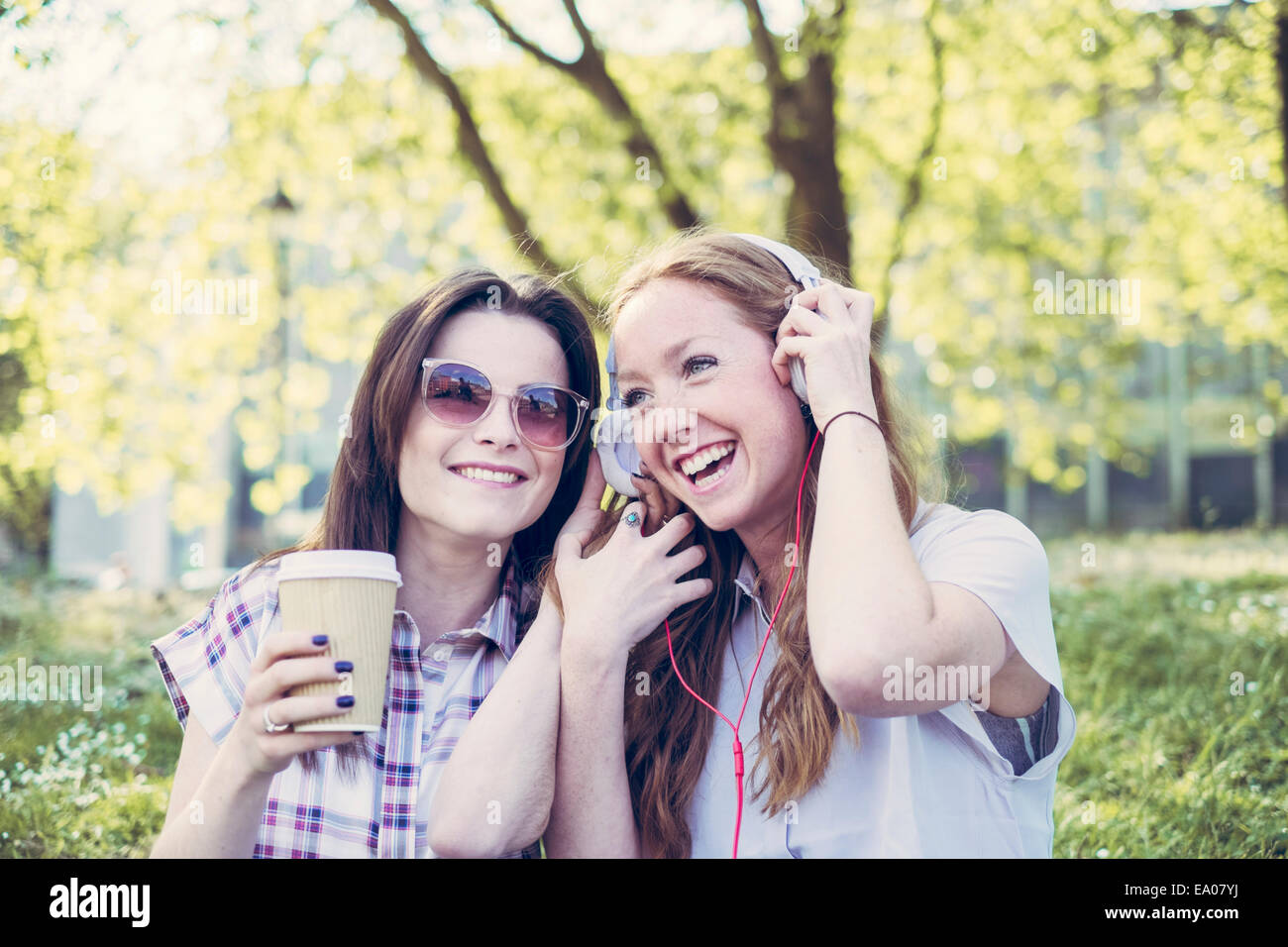 Two young female friends drinking coffee and listening to music on headphones in park Stock Photo