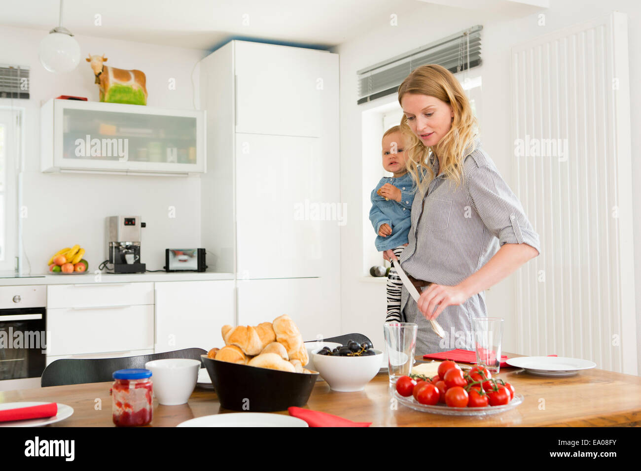 Mother setting kitchen table while holding baby daughter Stock Photo