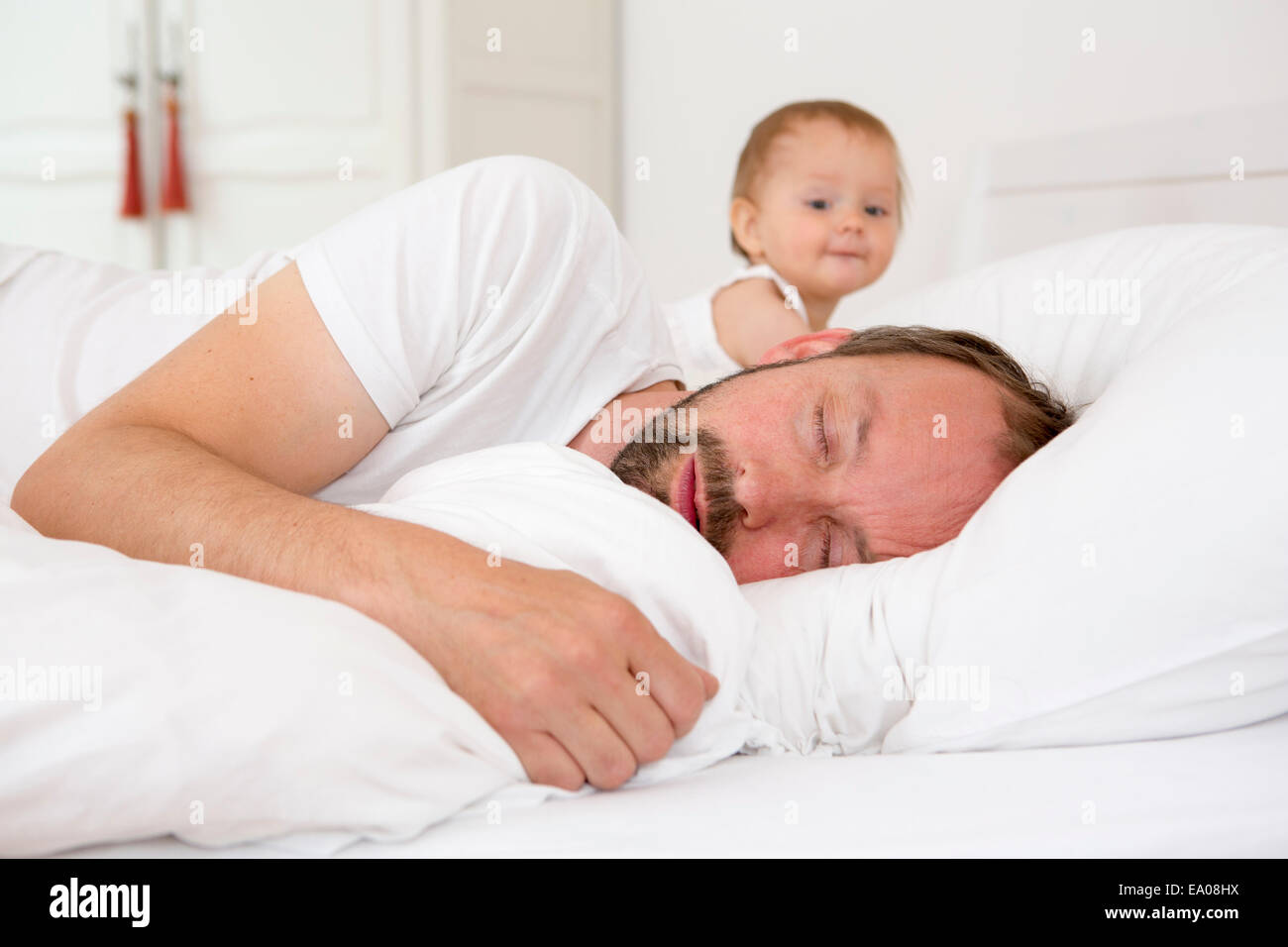 Father asleep in bed, baby daughter watching Stock Photo