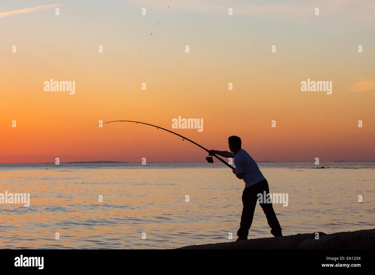 A lone angler on the shore sihouetted against an orange sunset Stock Photo