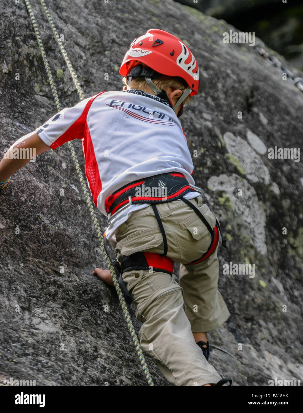 Boy child climbing a rock face in full climbing equipment looking down Stock Photo